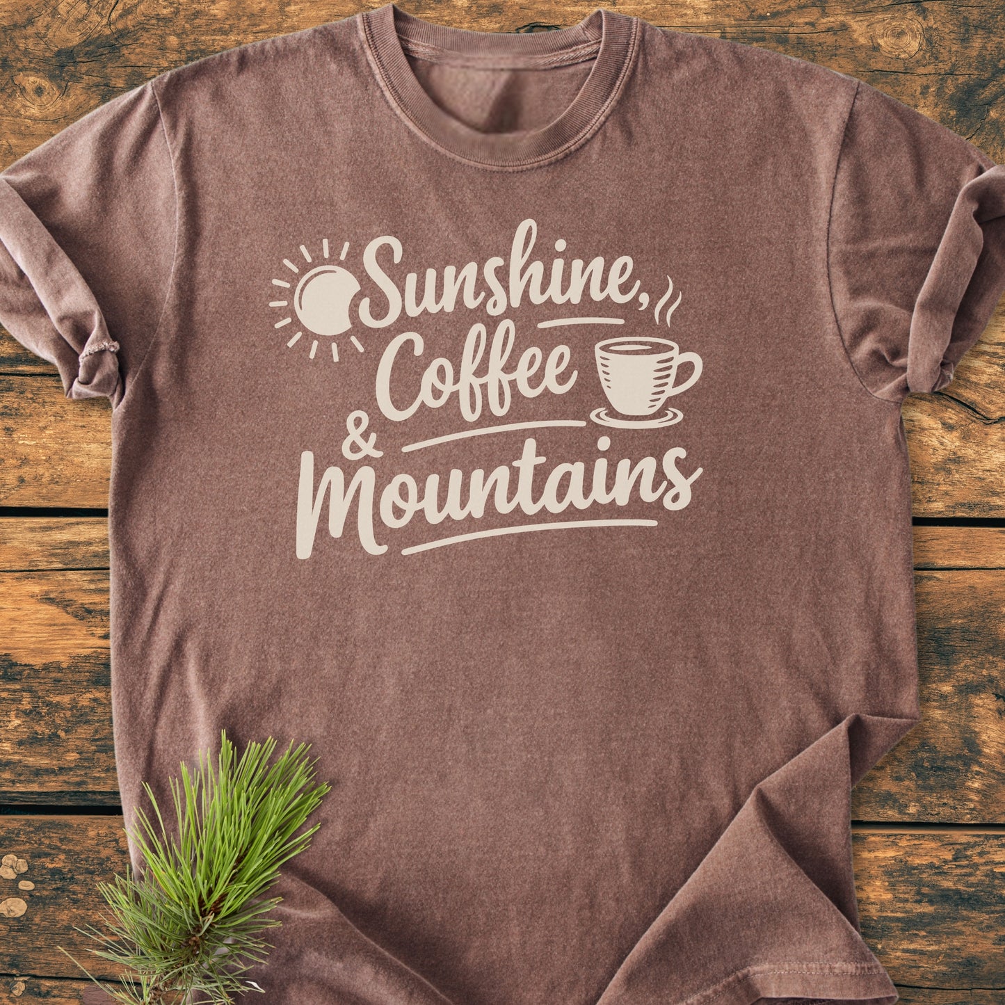 Sunshine, Coffee, Mountains - Vintage Heavyweight Tee featuring a phrase and icons printed in cream on a brown, short-sleeved shirt, laid on a wooden surface with pine needles for styling.