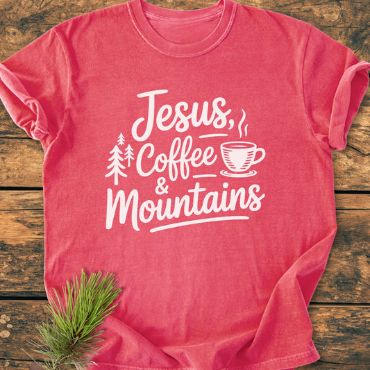 Jesus, Coffee, Mountains - Vintage Wash Tee features a white graphic with pine trees and coffee cup on a coral-red T-shirt, laid on wood with a pine sprig accentuating the outdoor theme.