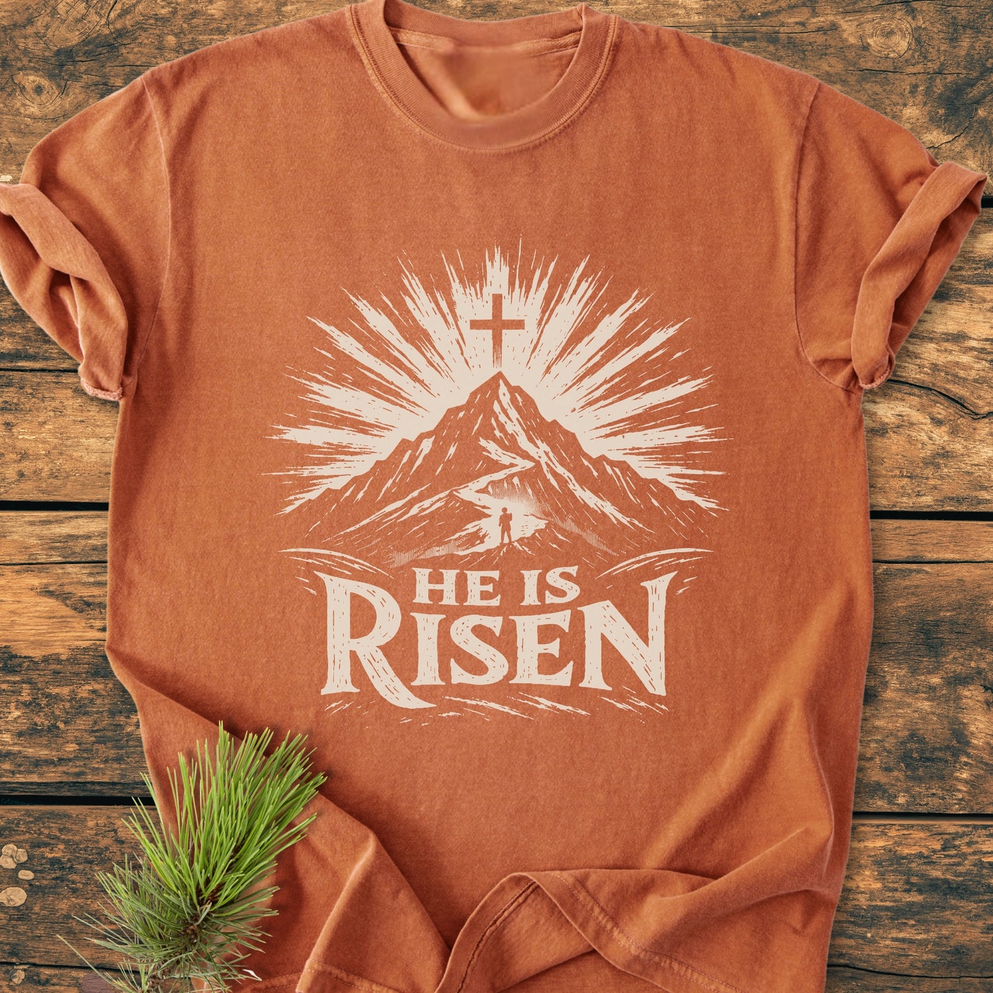 He Is Risen - Vintage Wash Tee features a stylized mountain sunrise design with a figure on a path, and 'HE IS RISEN' text, laid on wood with a pine sprig beside.