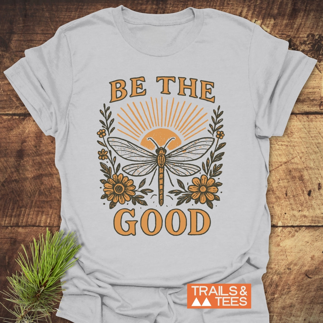Be The Good T-Shirt featuring a dragonfly with sun-ray lines, vintage-style BE THE GOOD text, and floral accents, laid on wood with a pine sprig, embodying nature-inspired comfort.