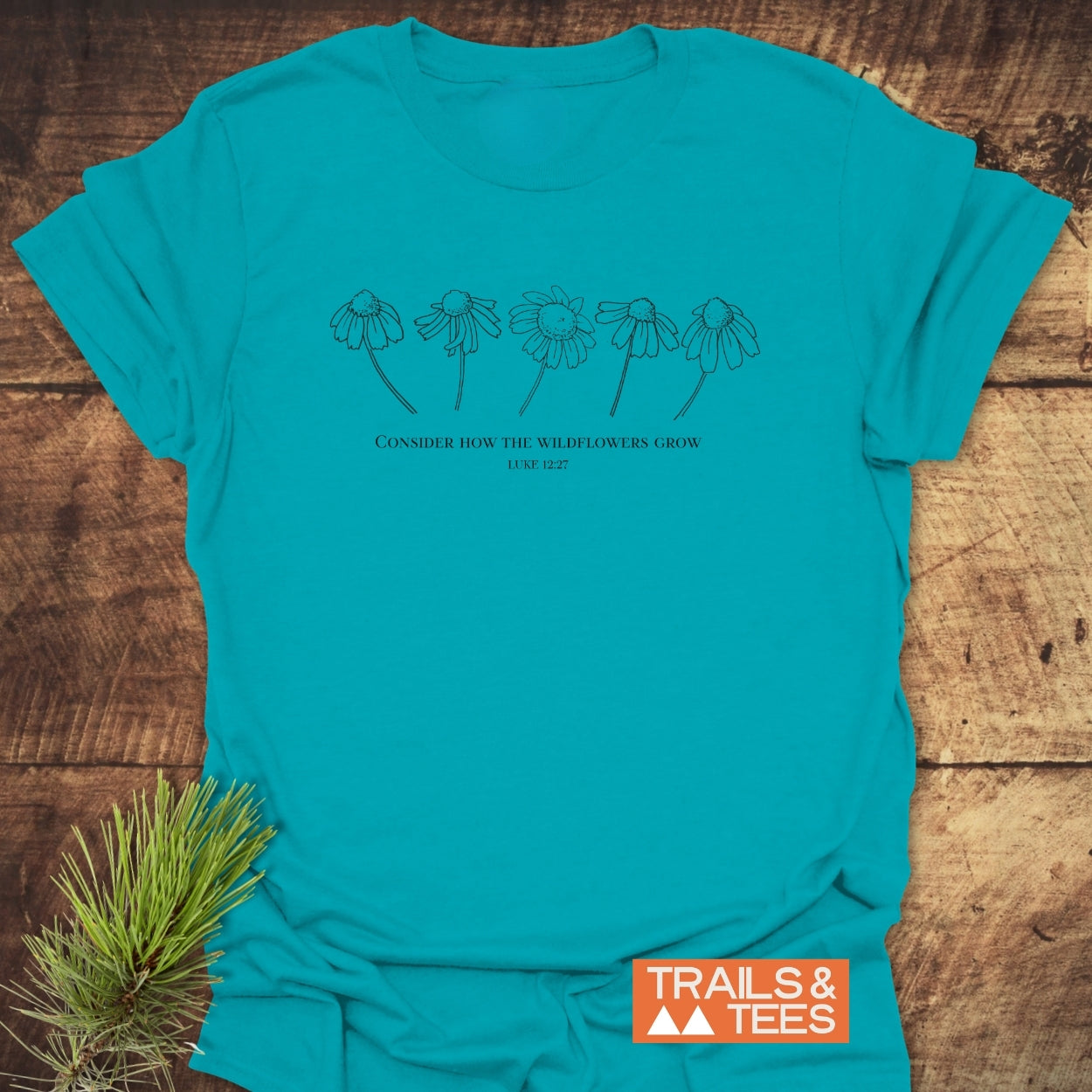 Consider How The Wildflowers Grow T-Shirt featuring five wildflower line drawings and a Luke 12:27 quote, displayed on a flat surface, showcasing its soft, durable cotton design and Trails & Tees logo.