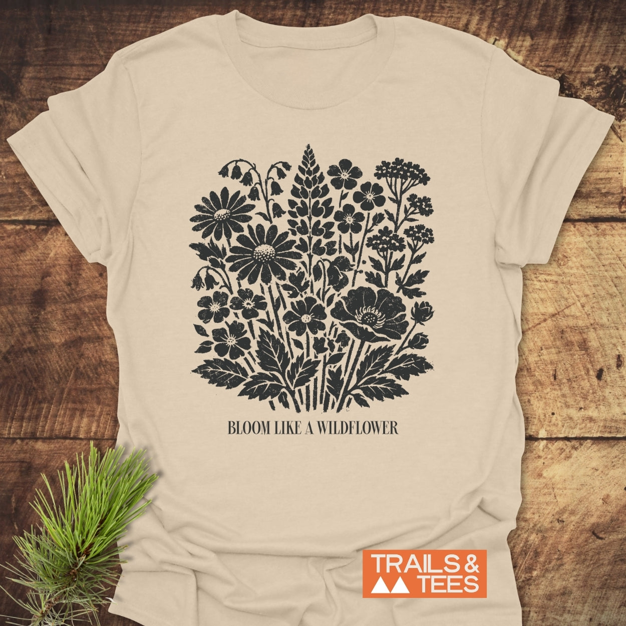 Bloom Like A Wildflower T-Shirt features a black wildflower silhouette with the slogan, laid flat on wood, demonstrating its rustic charm. Includes Trails & Tees logo, showcasing outdoor adventure spirit.
