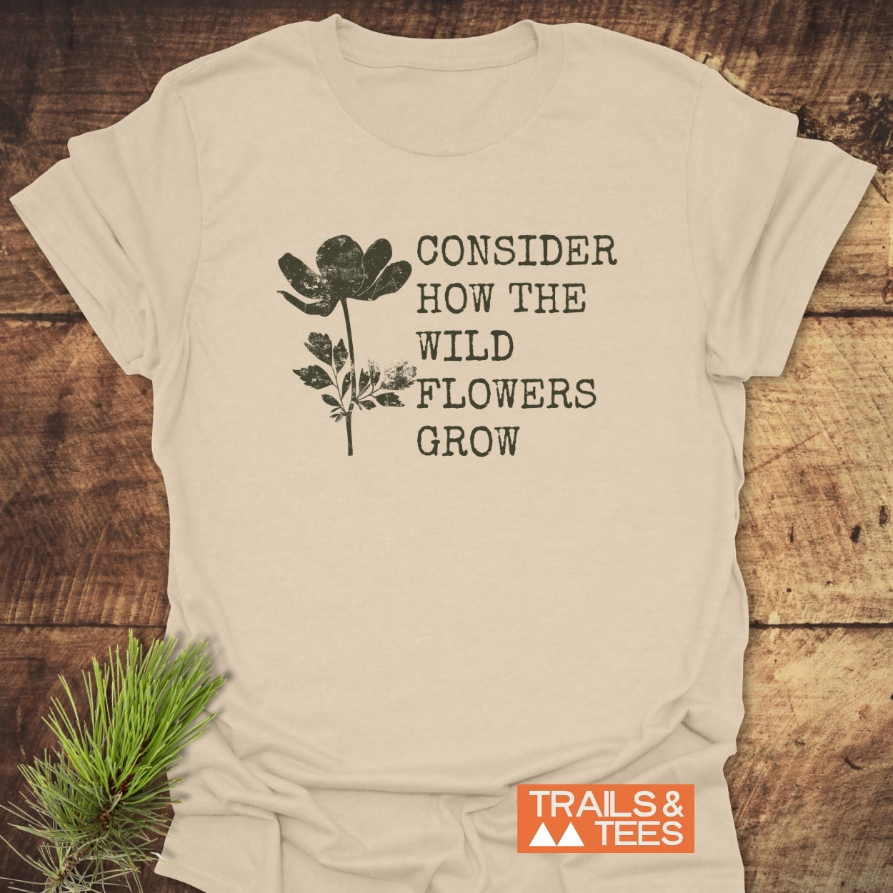 Consider How The Wild Flowers Grow T-Shirt, light-beige, features a black wildflower silhouette and phrase. Includes Trails & Tees logo. Made from soft, ring-spun cotton, pre-shrunk, durable print, and no side seams.