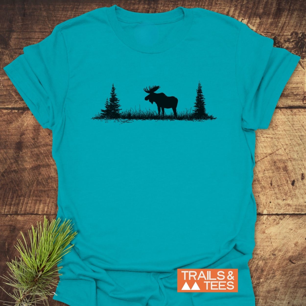 Lone Moose T-Shirt displayed flat, featuring a black moose and pine tree graphic. Styled with a pine branch, alongside the TRAILS & TEES logo, highlighting its outdoor adventure theme.
