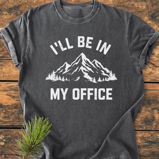 I'll Be In My Office - Vintage Wash Tee: Charcoal-gray T-shirt with a white mountain and tree graphic, laid on wood with pine bough, highlighting comfort and durability of garment-dyed ring-spun cotton.
