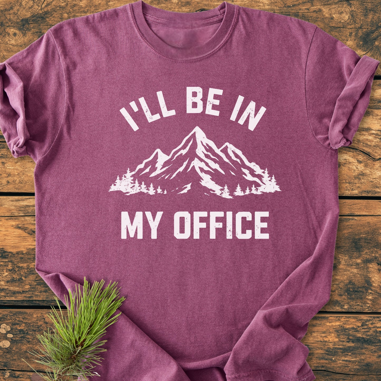 I'll Be In My Office - Vintage Wash Tee with a mountain range and pine trees graphic, folded on wood, featuring a pine sprig. Made from soft, durable 100% ring-spun cotton.