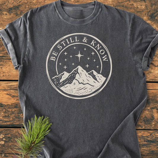 Be Still And Know, Psalm 46:10 - Vintage Wash Tee features a cream graphic of a mountain and starry sky, highlighting the phrase on a charcoal-gray Comfort Colors t-shirt laid flat.