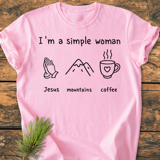 Pink I'm A Simple Woman. Jesus Mountains Coffee - Vintage Wash Tee on wooden surface, featuring icons of praying hands, mountains, and coffee mug. Made of soft, durable, pre-shrunk 100% ring-spun cotton.