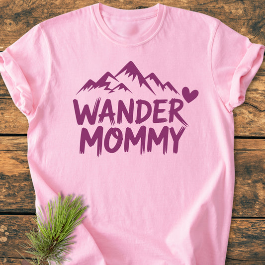 Wander Mommy - Vintage Heavyweight Tee: Pastel-pink T-shirt with purple mountain graphic and WANDER MOMMY text, laid on rustic wood. Suggests an outdoorsy theme with a pine sprig accent.