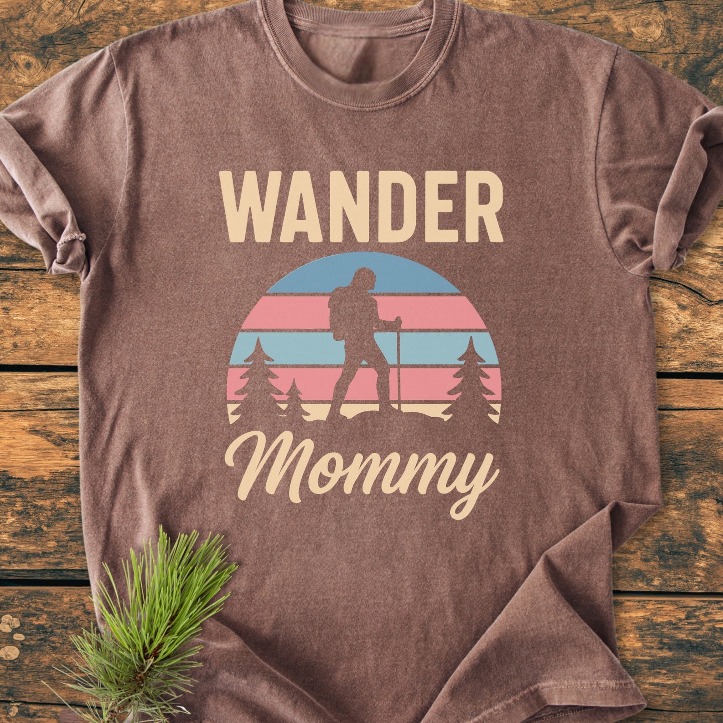 Wander Mommy Retro - Vintage Heavyweight Tee features a stylized sunset graphic with a hiker and pine trees, WANDER above, on soft, garment-dyed, brown fabric laid on wood.