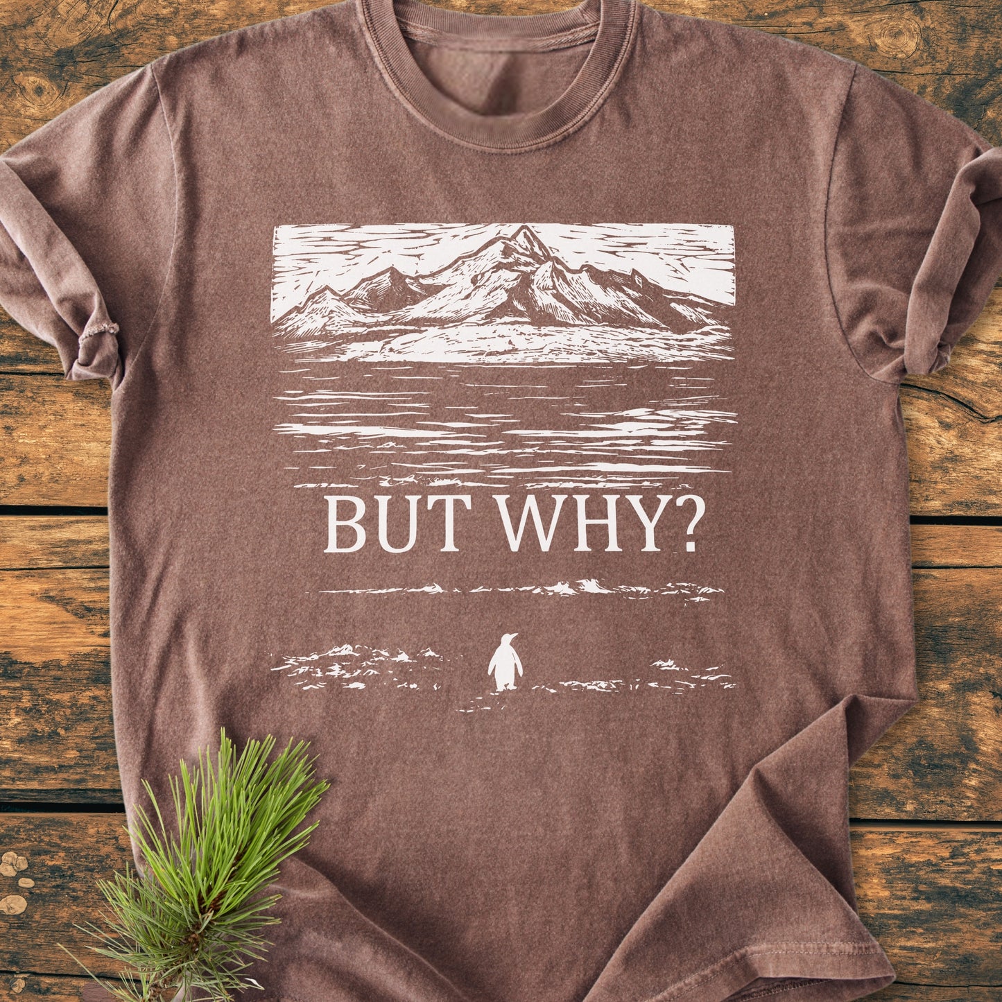 But Why - Vintage Heavyweight Tee features a white mountain landscape and penguin silhouette on a brown tee, lying flat with a pine sprig. Crafted from soft, durable, garment-dyed cotton for comfort.