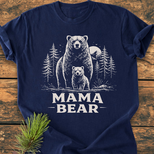 Mama Bear - Vintage Heavyweight Tee: Navy-blue T-shirt featuring a white forest scene with bears and mountains, MAMA BEAR text below. Laid on wood with a pine sprig beside it.