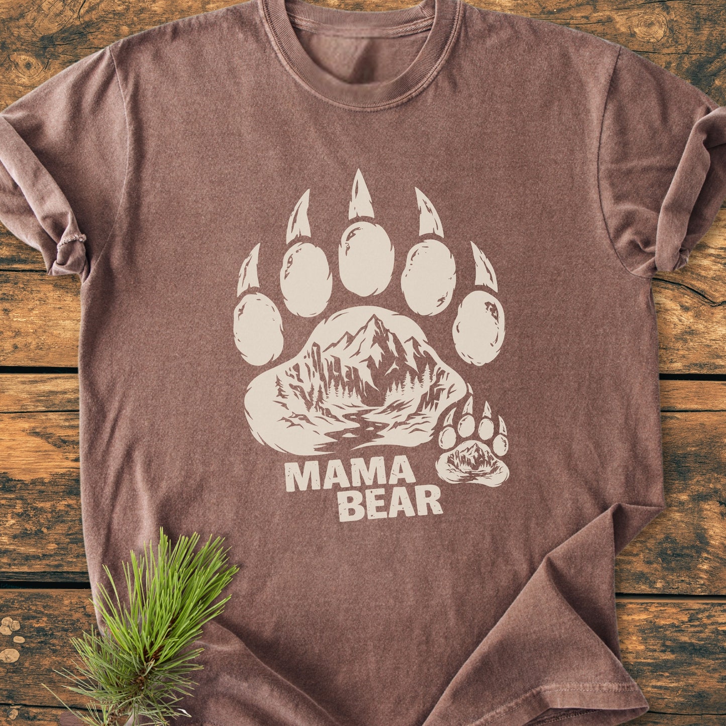 Mama Bear - Vintage Heavyweight Tee featuring a large bear paw print with mountain scenes on a brown, short-sleeve shirt, laid on a wooden surface, complemented by a pine branch for an outdoorsy vibe.