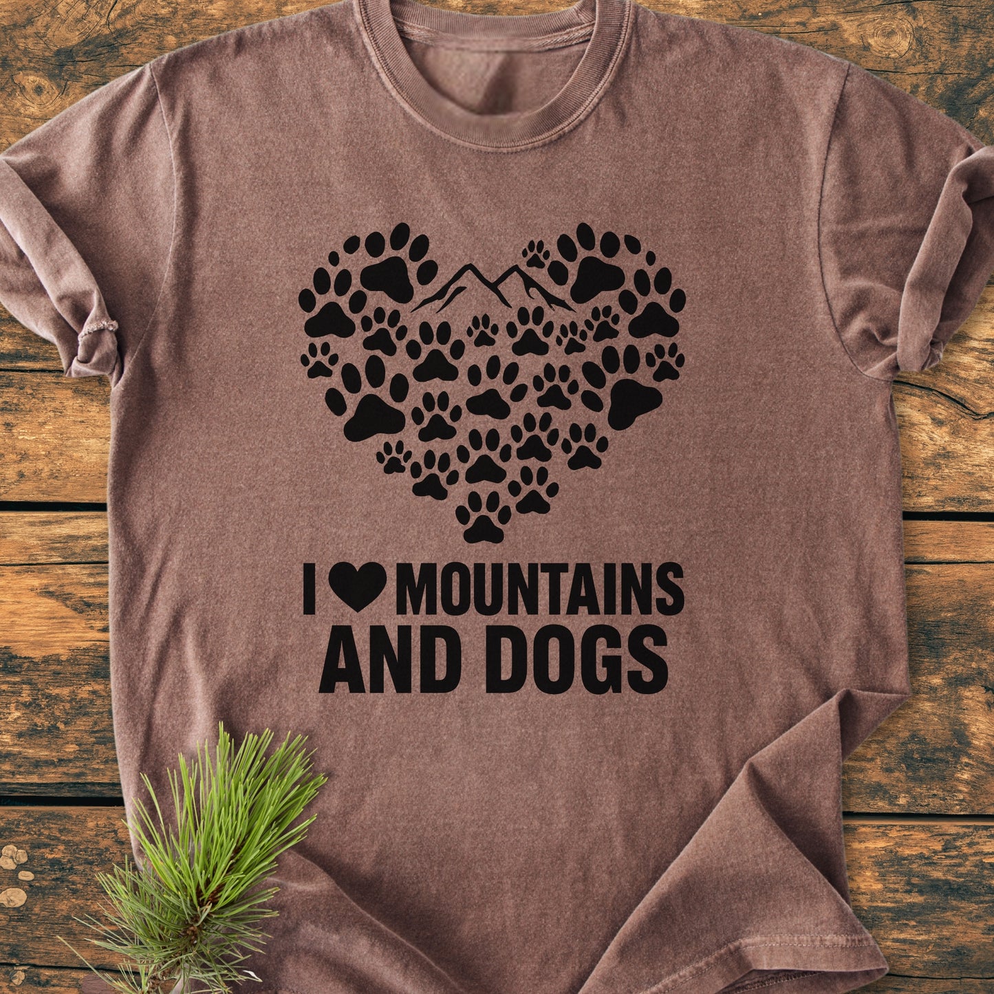 Paw Heart Dog Lover - Vintage Heavyweight Tee features a heart graphic of paw prints and mountains, with I ♥ MOUNTAINS AND DOGS text, displayed on a wooden surface with a pine sprig.