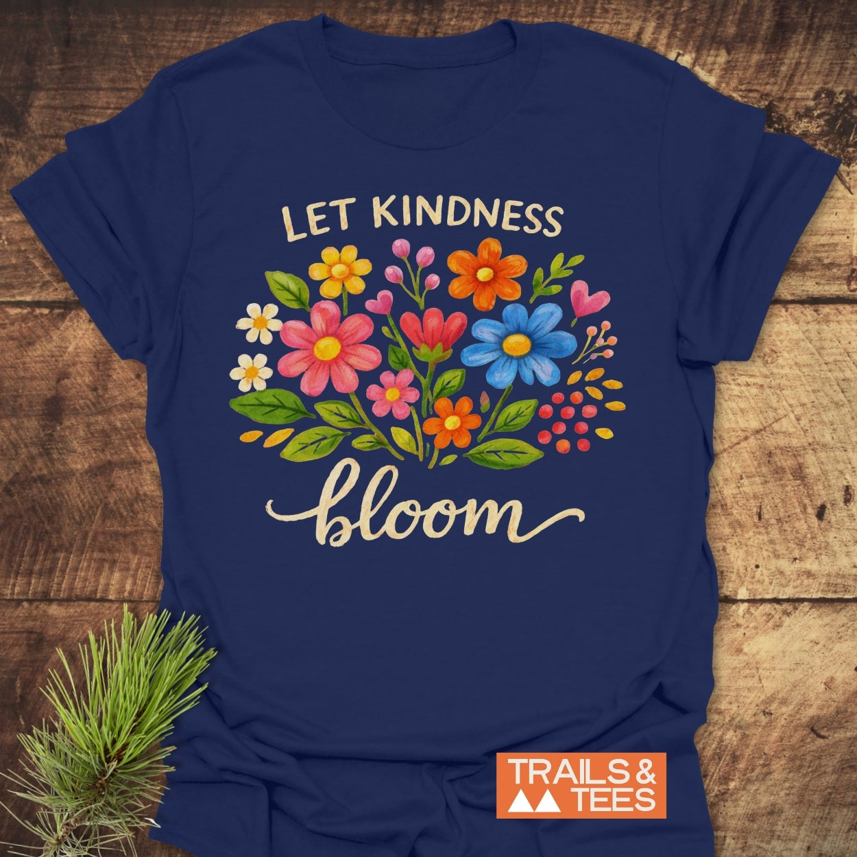 Let Kindness Bloom T-Shirt features a hand-painted floral bouquet with Let Kindness bloom text. Made from soft, ring-spun cotton, it includes a small TRAILS & TEES logo on the front.