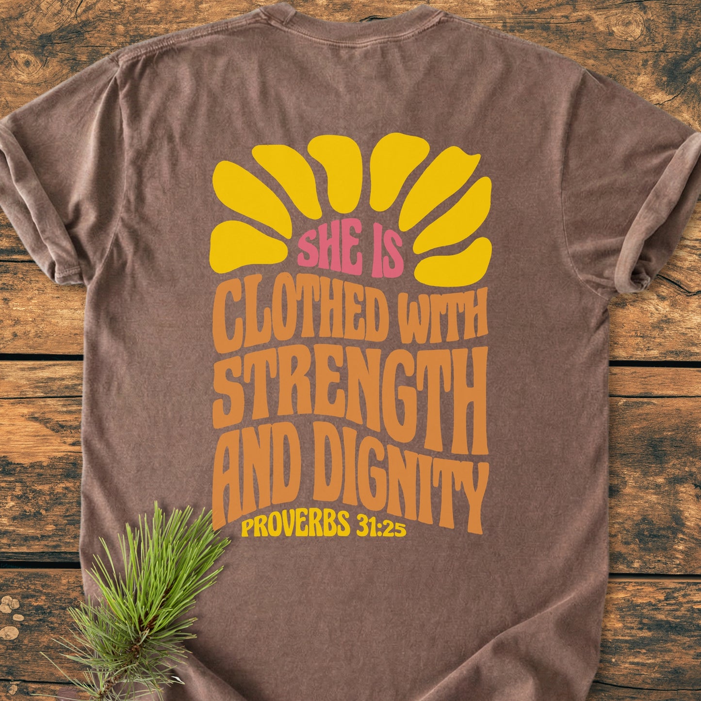 Strength And Dignity BACK PRINT - Vintage Wash Tee on wooden surface, featuring yellow sun-ray graphic and Proverbs 31:25 text in groovy lettering, tucked pine sprig under corner for rustic touch.