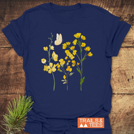 Yellow Boho Flowers T-Shirt featuring stylized wildflowers and butterfly graphic on navy. Laid flat on wood, with pine needle accent; Trails & Tees brand watermark visible.