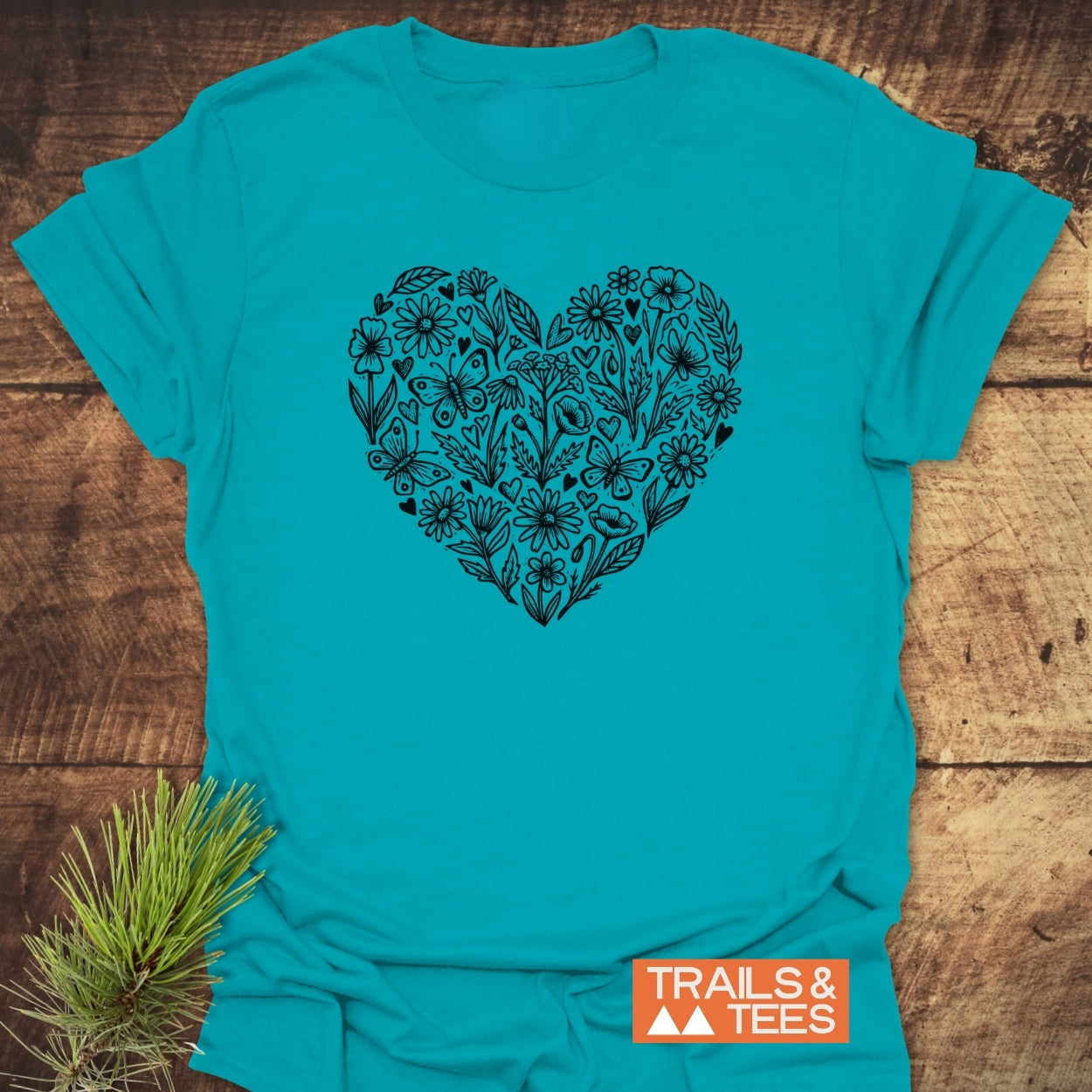 Wildflower Heart Butterfly Soul Softstyle T-Shirt featuring a large black heart of doodled flora and fauna on teal, laid against wood, with TRAILS & TEES logo and pine sprig accentuating its outdoor essence.