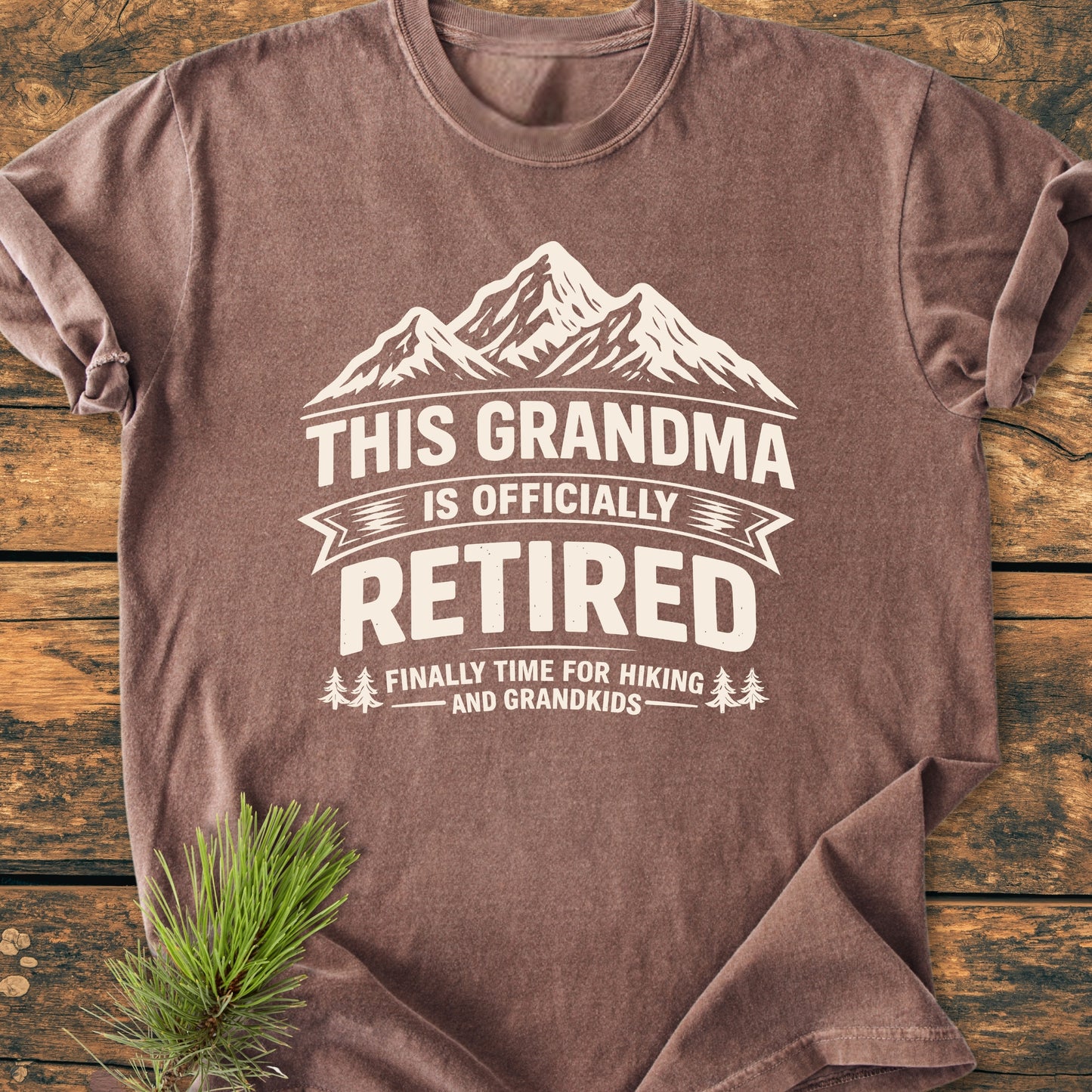 This Grandma is Officially Retired - Vintage Wash Tee features a heathered brown finish with a white mountain graphic and playful retirement message, laid on wood with a pine sprig accent.