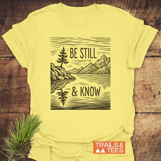 Be Still And Know T-Shirt featuring a lakeside scene illustration with a pine tree and mountains, styled with a pine sprig. Part of Trails & Tees collection, combining faith and nature.