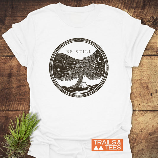 Be Still Softstyle T-Shirt featuring a circular black line-art of a pine tree under a starry sky with crescent moon, a tent, and the words BE STILL above.
