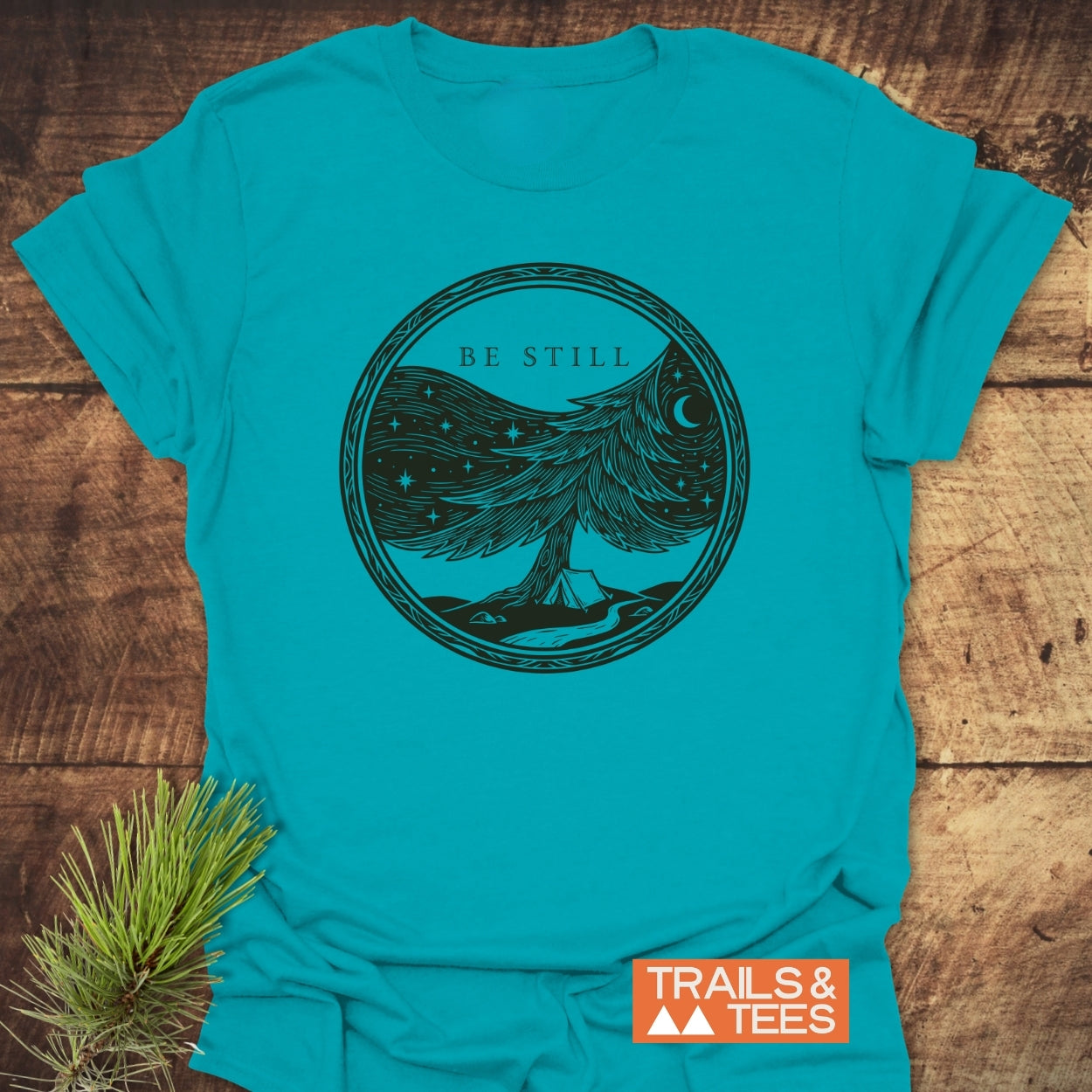 Be Still Softstyle T-Shirt with a black line-art design featuring BE STILL, a starry tree, tent, and stream. Branded with the TRAILS & TEES logo, laid flat beside a pine sprig.
