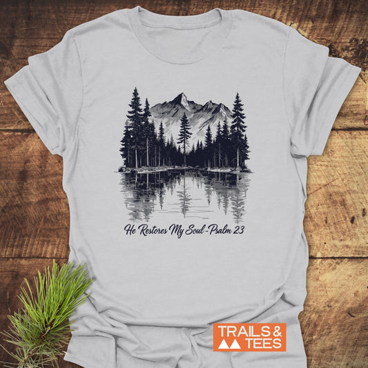 Light-gray He Restores My Soul T-Shirt featuring a mountain and pine tree illustration, Psalm 23 quote, and Trails & Tees logo, laid on wood with pine sprigs nearby.