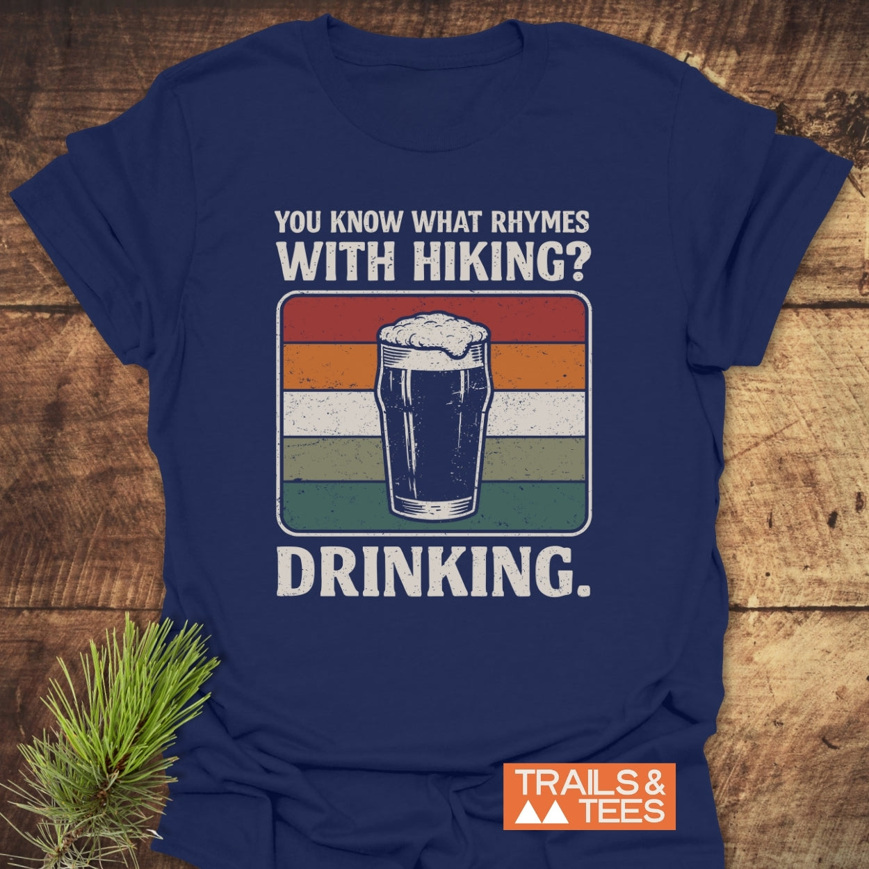 What Rhymes With Hiking? Softstyle T-Shirt featuring a retro-style beer graphic and humorous text, laid flat on wood with a pine sprig, perfect for outdoor enthusiasts who enjoy post-hike drinks.