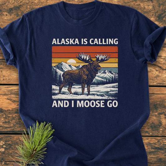 Alaska Is Calling - Vintage Wash Tee featuring a retro bull moose illustration in a snowy landscape with a sunset sky, paired with a punny slogan, laid on a wooden surface.