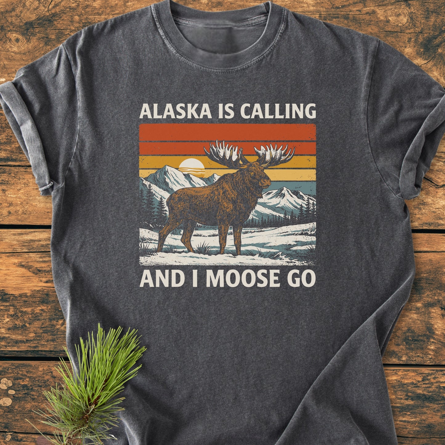 Alaska Is Calling - Vintage Wash Tee featuring a retro moose graphic in a snowy landscape with sunset, laid on wood. Beside it, a pine sprig enhances the nature-inspired theme.