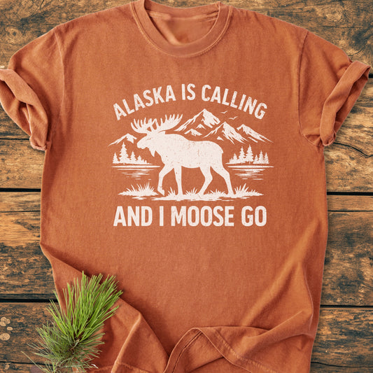 Alaska Is Calling - Vintage Wash Tee features a white moose graphic with mountains and forest on a rust-orange shirt, styled with a pine sprig, showcasing a relaxed fit and vintage wash.