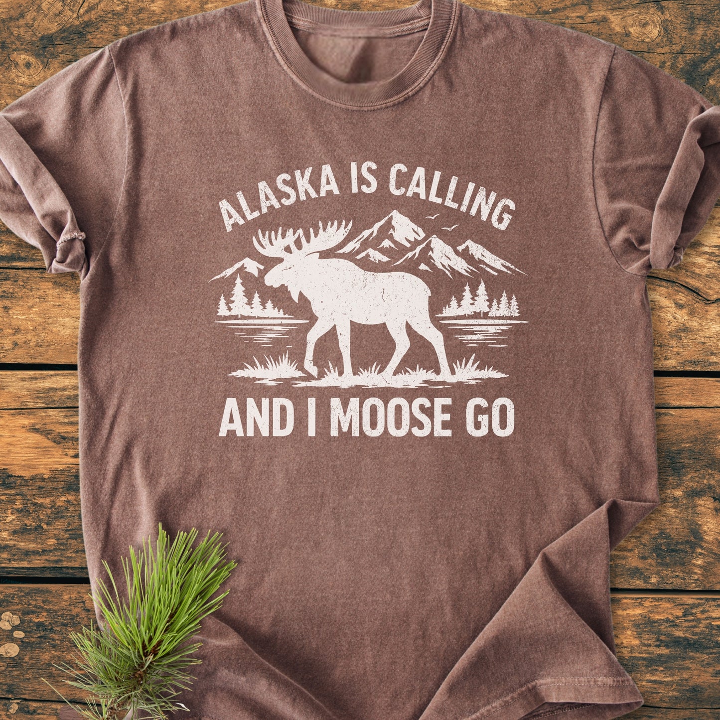 The Alaska Is Calling - Vintage Wash Tee features a moose silhouette against mountains and pines, with playful text. Laid on wood, it embodies a relaxed, soft-washed vintage style.