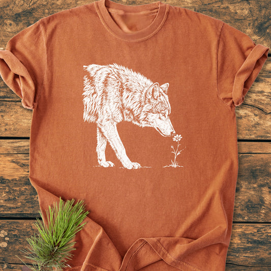 Stop And Smell The Flowers - Vintage Wash Tee featuring a white wolf sniffing a flower, styled with a pine sprig under the sleeve, laid on a wooden background.