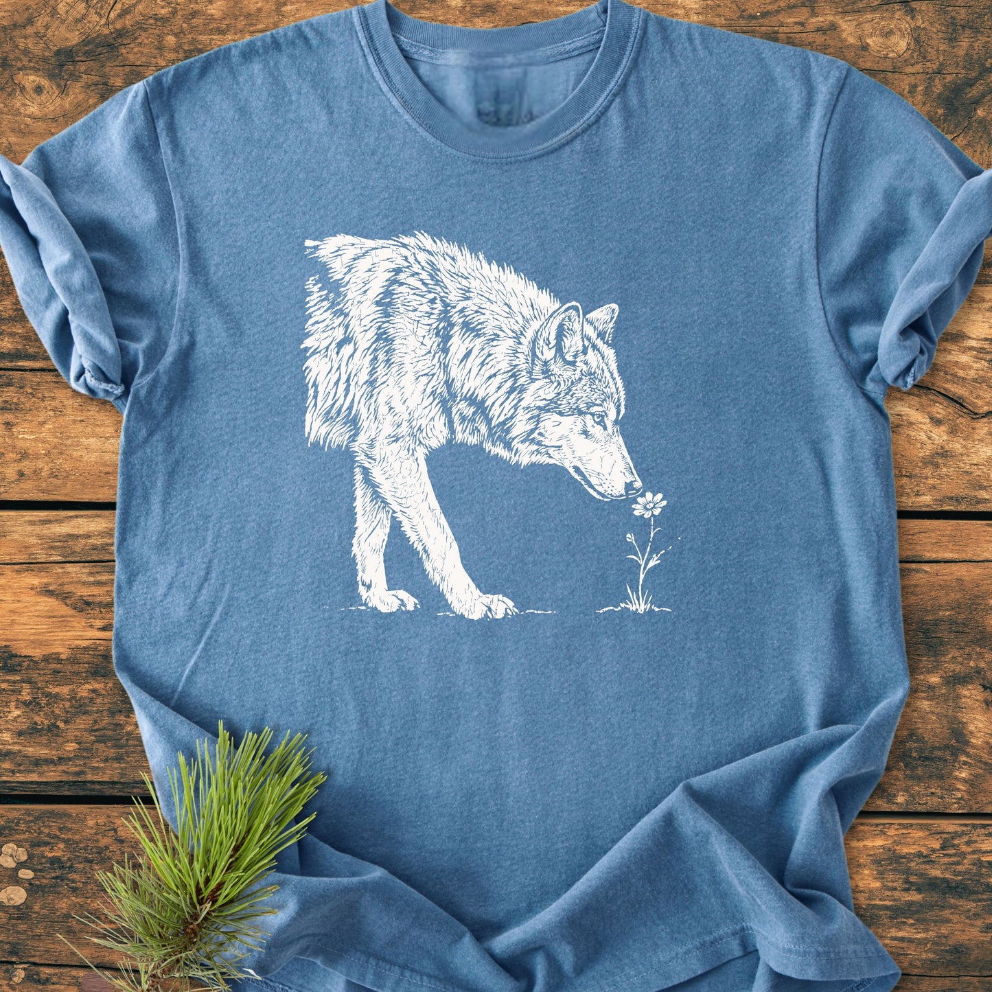 Stop And Smell The Flowers - Vintage Wash Tee featuring a screen-printed wolf sniffing a flower, laid flat on wood with a pine sprig tucked under the corner for a nature-inspired look.