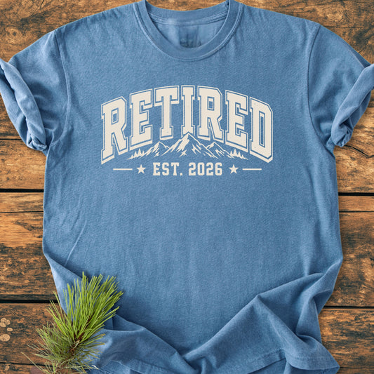 Retired 2026 - Vintage Wash Tee in dusty-blue with RETIRED collegiate graphic, mountain range, and EST. 2026. Laid on a wooden surface with pine sprig accentuating its outdoor theme.
