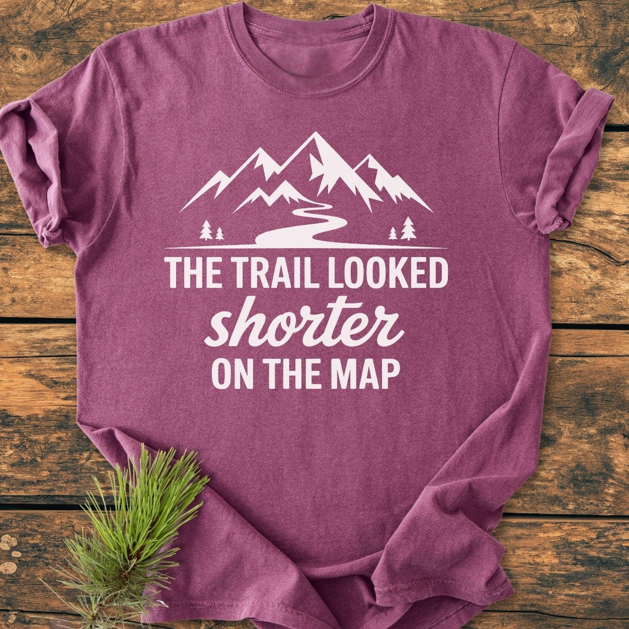 Heather-purple T-shirt featuring stylized mountains, trail, and trees graphic with slogan THE TRAIL LOOKED shorter ON THE MAP, laid on wood. Ideal for hikers, vintage wash, unisex fit.