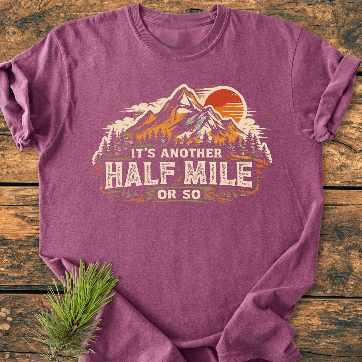 Another Half Mile Or So - Vintage Wash Tee featuring a mountain scene with snow peaks, trees, and sun. Laid on wood, complemented by a pine sprig, embodying hiking adventures and outdoor spirit.
