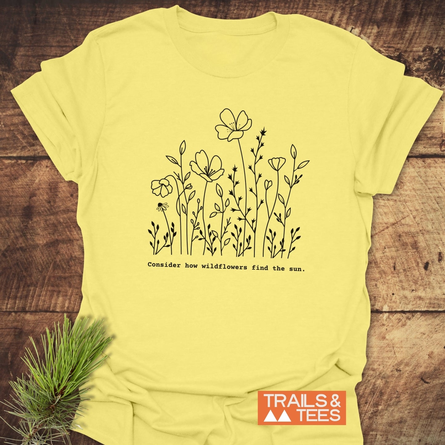 How Wild Flowers Find The Sun T-Shirt with a black wildflower illustration and quote. Features an orange TRAILS & TEES logo, displayed on wood with a pine sprig beside.