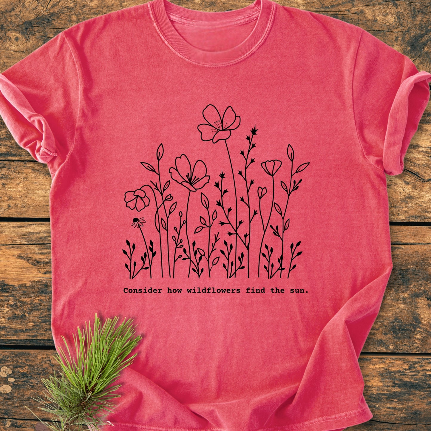 How Wildflowers Find The Sun - Vintage Wash Tee features a black wildflower illustration with text on a reddish-pink shirt, flat-laid on wood with a pine sprig, embodying nature-inspired elegance.