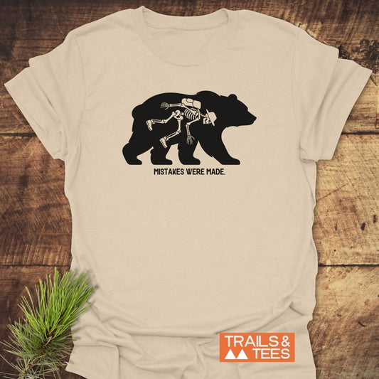 Mistakes Were Made T-Shirt featuring a bear silhouette with a skeleton hiker inside, captioned below. Light-tan tee displayed on wood, adorned with a pine sprig, and Trails & Tees logo visible.