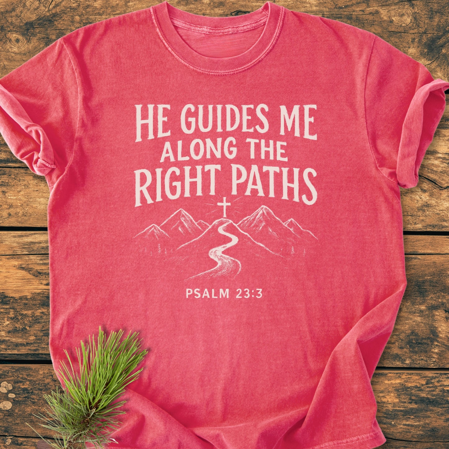 He Guides Me - Vintage Wash Tee featuring a hand-drawn graphic of a cross, mountain range, and Psalm 23:3 text, laid flat on wood with pine needles, illustrating faith and nature.
