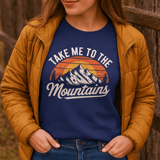 Person wearing Take Me To The Mountains T-shirt with mountain graphic and slogan, layered under a quilted jacket, hands in pockets.
