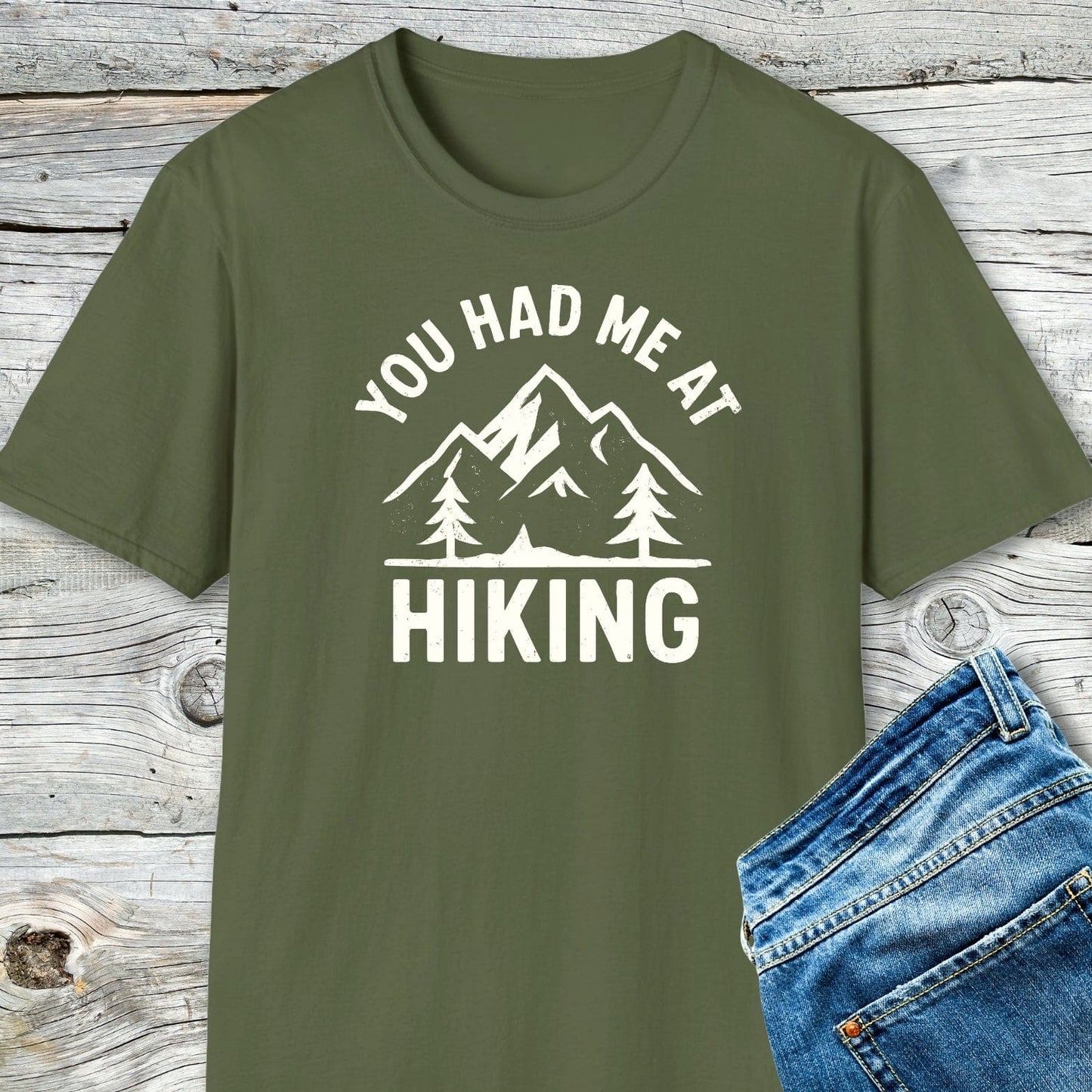 You Had Me At Hiking T-Shirt featuring mountain graphic and slogan, laid on wooden surface. Made of soft cotton, perfect for outdoor enthusiasts.