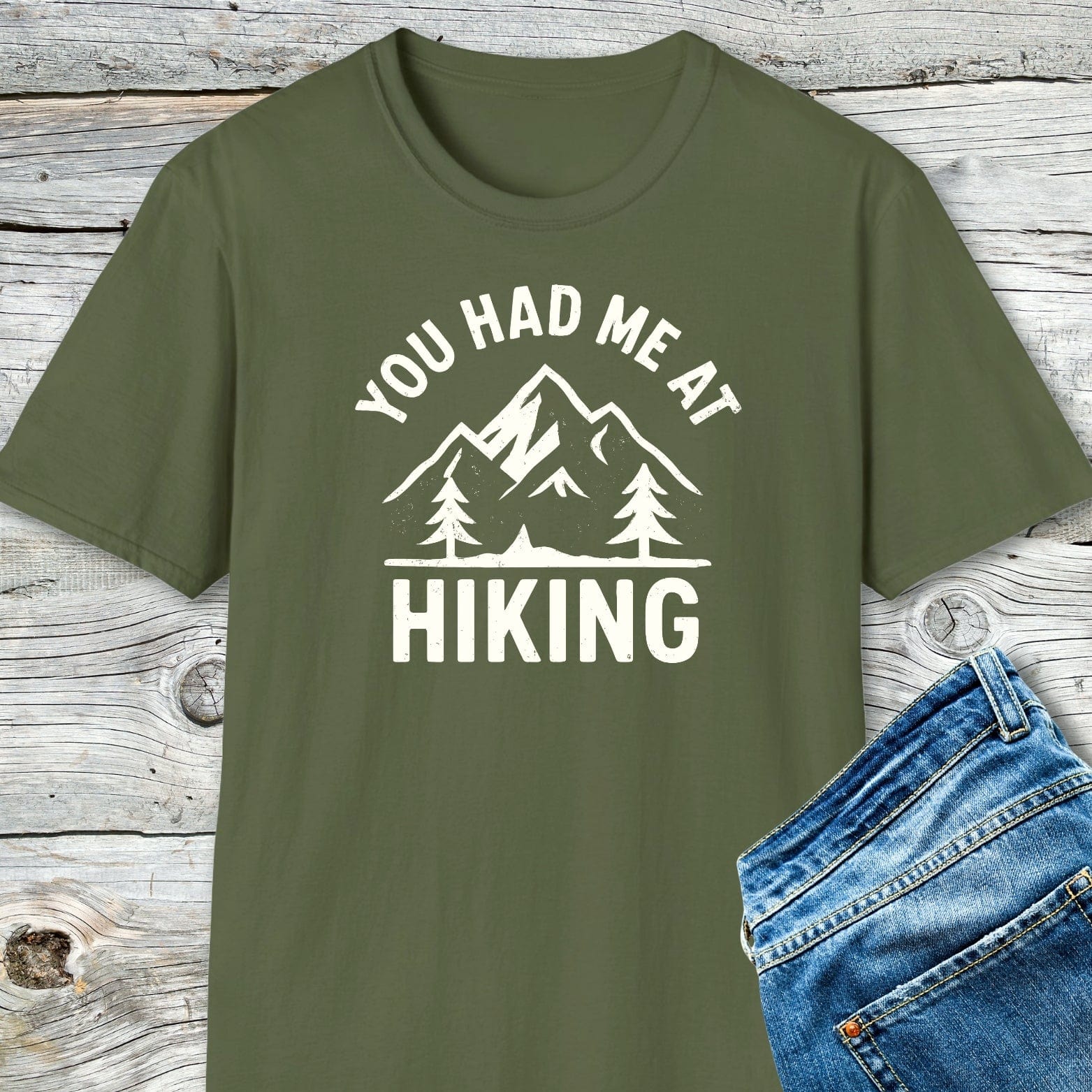 You Had Me At Hiking T-Shirt featuring mountain graphic and slogan, laid on wooden surface. Made of soft cotton, perfect for outdoor enthusiasts.