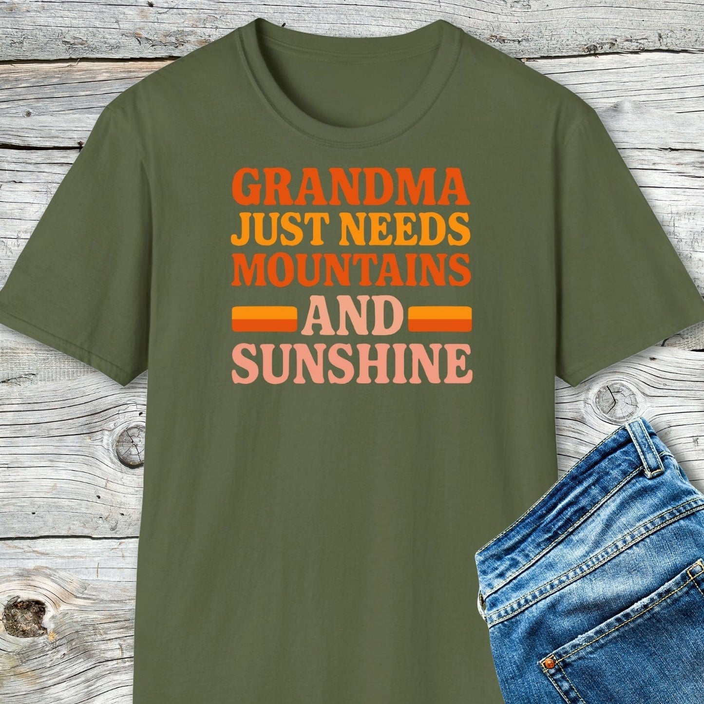 Retro 80s Grandma Mountains T-Shirt with bold lettering, laid flat on wood. Paired with blue denim jeans, showcasing outdoor adventure style and comfort.
