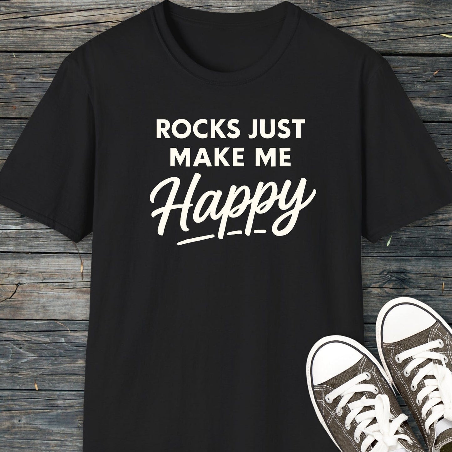 Black Rocks Make Me Happy T-shirt on wood surface, paired with gray sneakers. Lightweight, ring-spun cotton, durable print, pre-shrunk fit.
