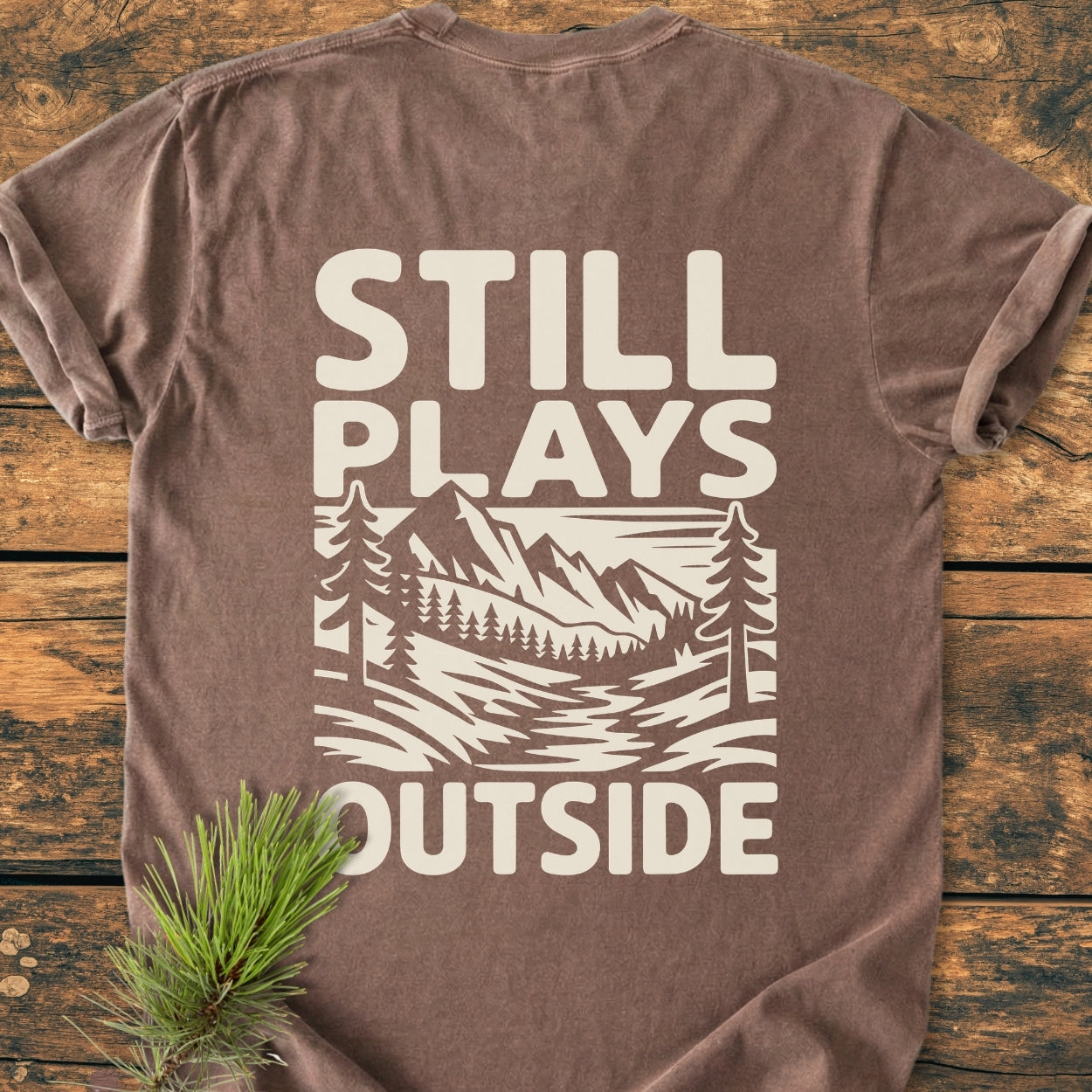 Still Plays Outside BACK PRINT - Vintage Wash Tee features a cream print of mountains, trees, and stream on a brown short-sleeve shirt, laid flat on a wooden surface with pine sprig accent.