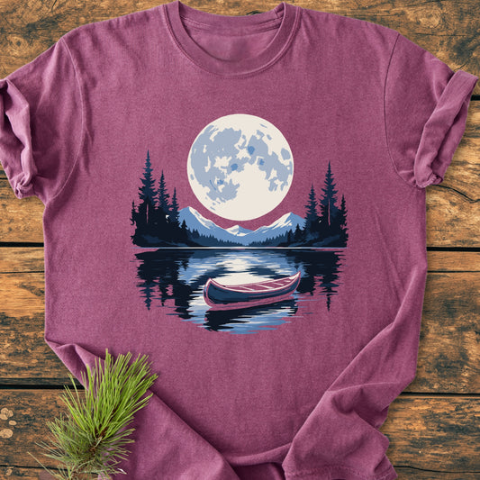 Moonlit Canoe - Vintage Wash Tee displayed on a wooden surface, features a full moon over a lake with a canoe, surrounded by mountains and pine trees, enhancing its rustic charm.
