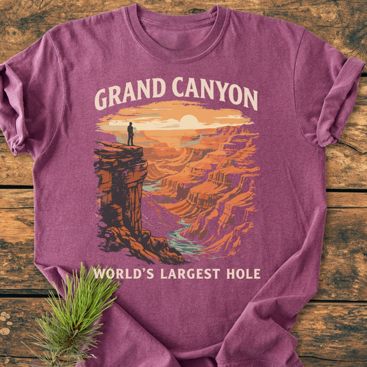 Grand Canyon Large Hole - Vintage Wash Tee on wood, featuring a retro Grand Canyon print at sunrise/sunset with a lone figure. Adjacent is a pine sprig, emphasizing its nature-inspired theme.