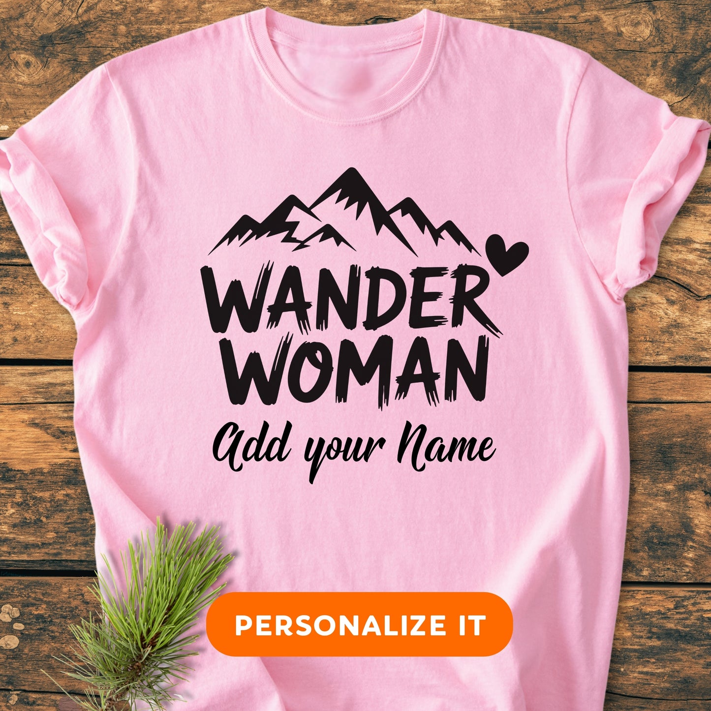 Wander Woman | Custom Name | Vintage Wash Tee featuring soft pink fabric with WANDER WOMAN text, mountain graphic, and personalization option. Laid on wood with pine sprig accent, includes PERSONALIZE IT button.